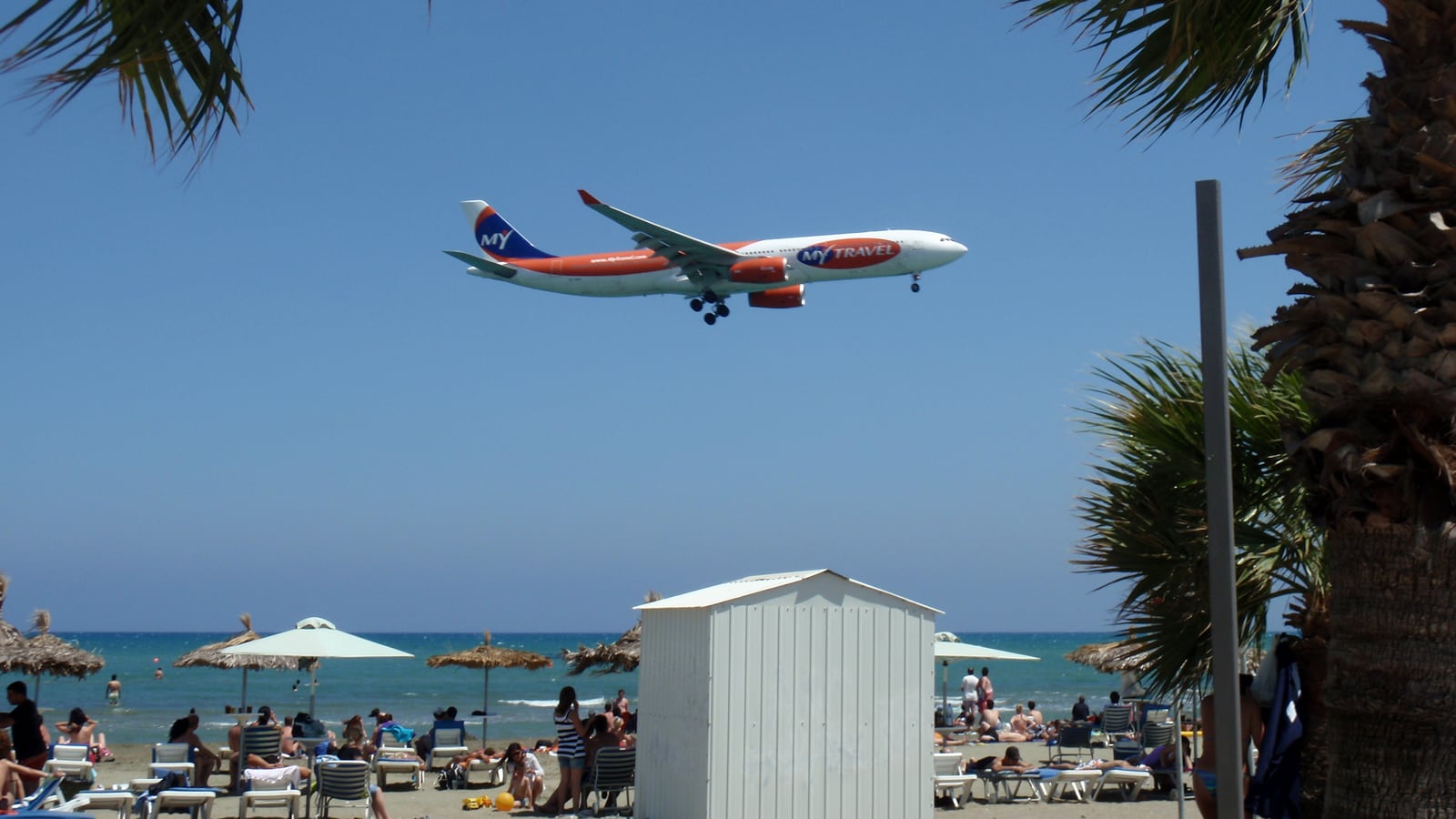 Avión de pasajeros volando bajo sobre una playa con sombrillas